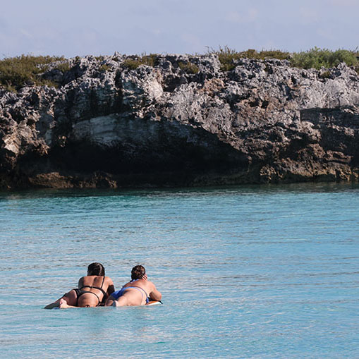 Two women floating peacefully in crystal-clear turquoise waters near rocky limestone formations in The Bahamas, demonstrating the pristine destinations and exceptional water clarity that guests experience on Breaking Free Sailing charters