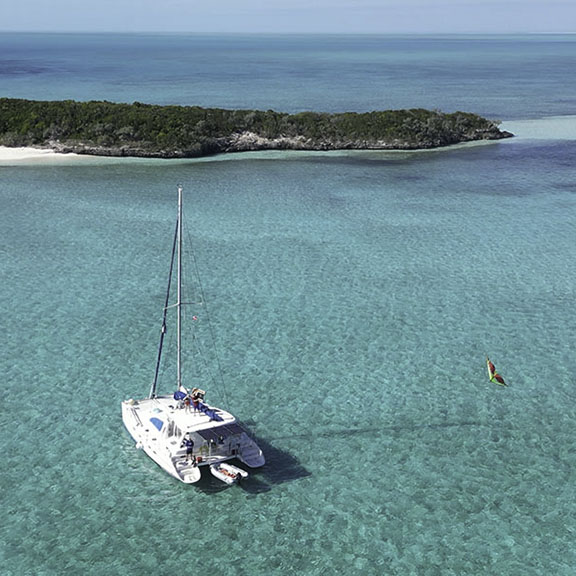 Guests exploring remote white sand beach surrounded by turquoise waters with dramatic sky and boats anchored in background - demonstrating the secluded paradise destinations accessible only through Breaking Free Sailing adventures