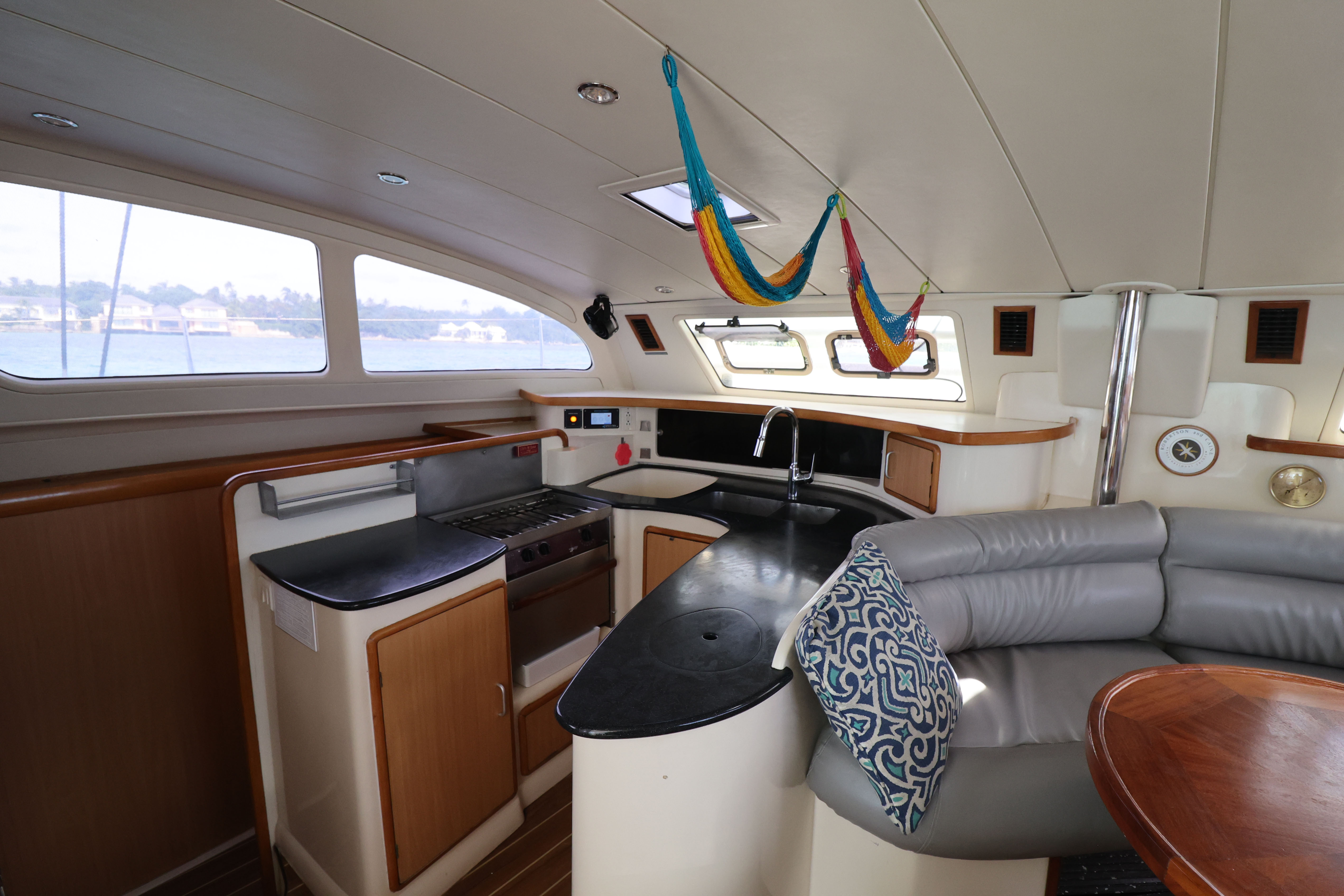 Interior view of Bon Bida catamaran galley showing curved seating with blue patterned cushions, dark countertops, compact kitchen with sink and stove, colorful hammocks hanging from ceiling, and large windows providing natural light - demonstrating the authentic, lived-in character of our sailing vessel where all meals are prepared