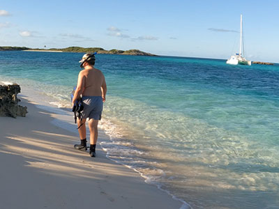 Person walking on pristine white sand beach with crystal clear turquoise water and sailboat in background in the Exuma Islands