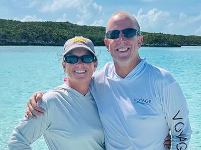Rob and Emily, your hosts and crew of Breaking Free Sailing, smiling together in front of beautiful turquoise Bahamian waters
